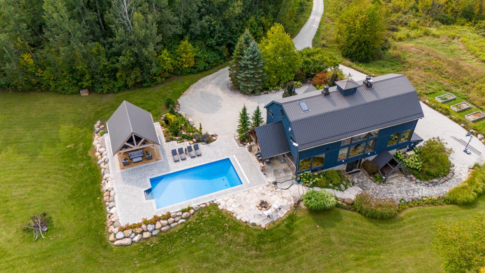 This image shows an arial photo of a two-story house with an outdoor swimming pool, lounge chairs, a pergola with covered patio, and with a firepit and a boulder retaining wall.