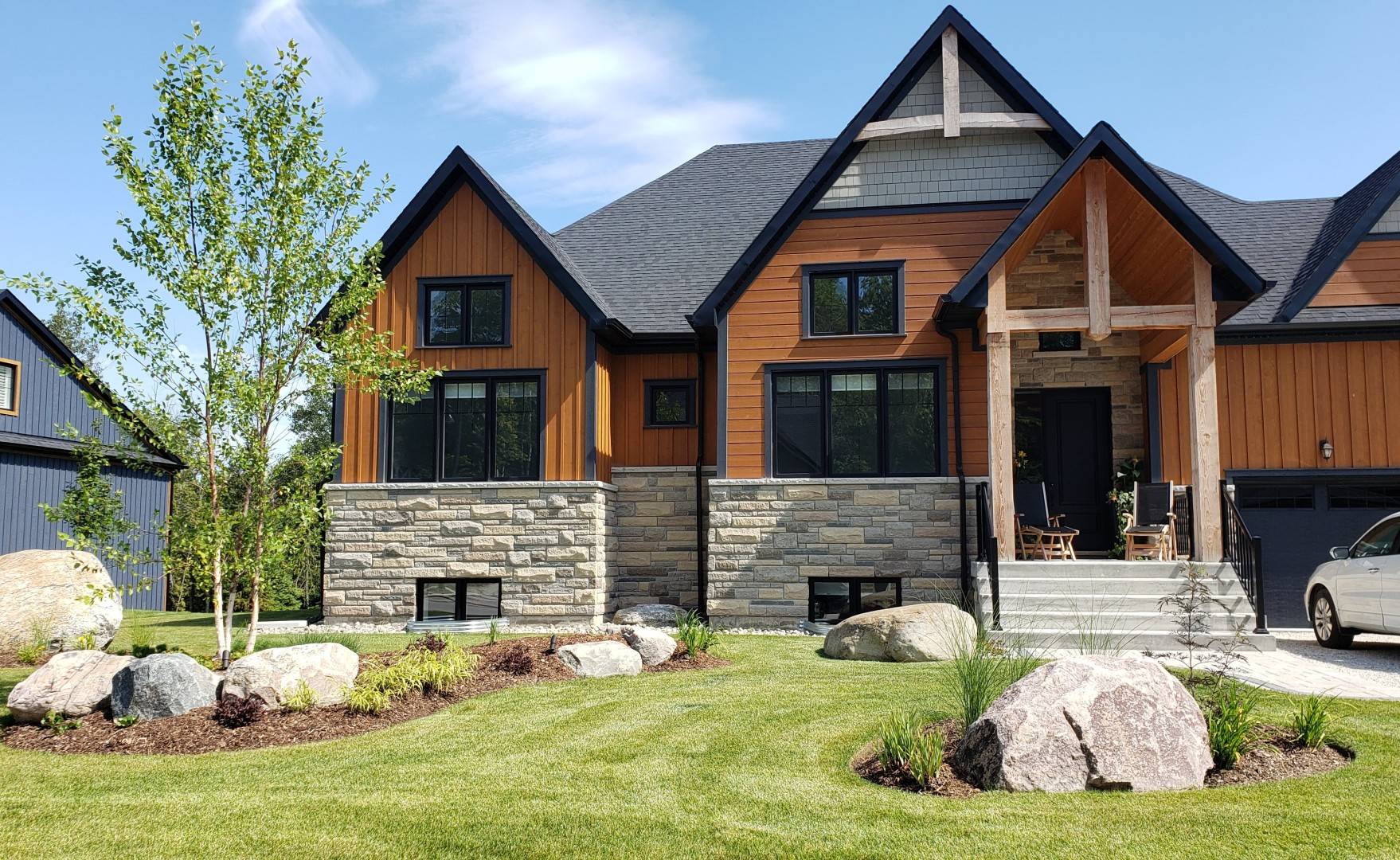 Modern home with a mix of wood and stone exterior, surrounded by a neatly landscaped front yard featuring boulders, young trees, and fresh mulch beds under a clear blue sky.