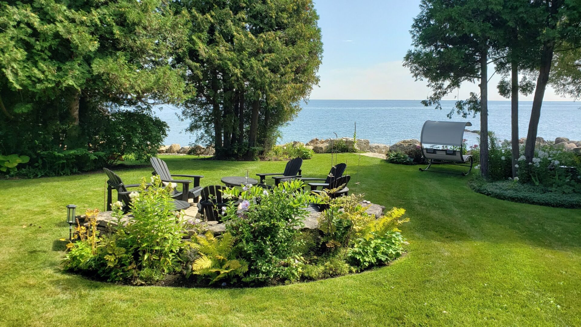 Lakeside backyard with a circular stone patio surrounded by Adirondack chairs and flowering garden beds, set on a manicured lawn with tall trees, a curved garden swing, and a clear view of the calm blue water beyond.