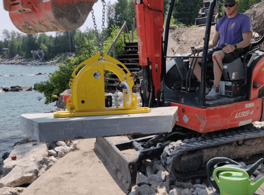 Man operating a red mini excavator places a large concrete slab using a yellow hydraulic clamp attachment beside a rocky shoreline, with calm blue water and forested waterfront properties in the background.