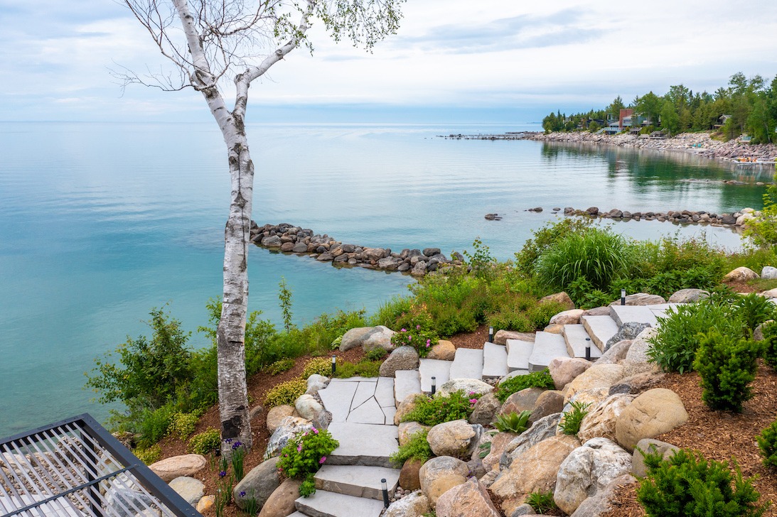 Stone steps wind through a landscaped garden with boulders, flowering plants, and a white birch tree, leading down to the calm, turquoise waters of a lakeshore bordered by a rocky breakwater and distant waterfront homes.